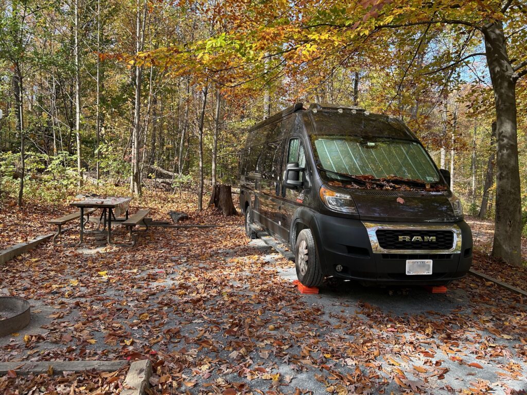  A charcoal gray camper van parked on a sloped gravel pad. There are trees beside and behind it in autumn colors with many leaves on the ground and collected on the van's windsheild.