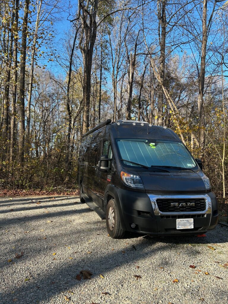 A charcoal gray camper van parked on a gravel camp site with tall trees behind and right of it.