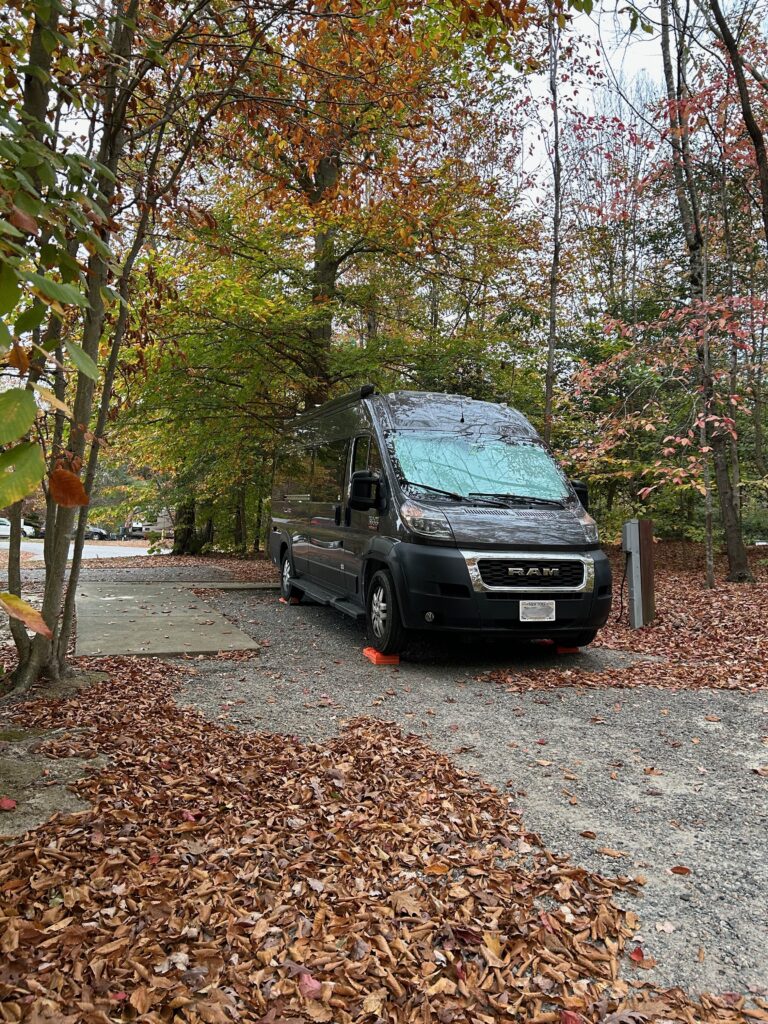 Camper van parked on a gravel campsite with trees and autumn leaves all around